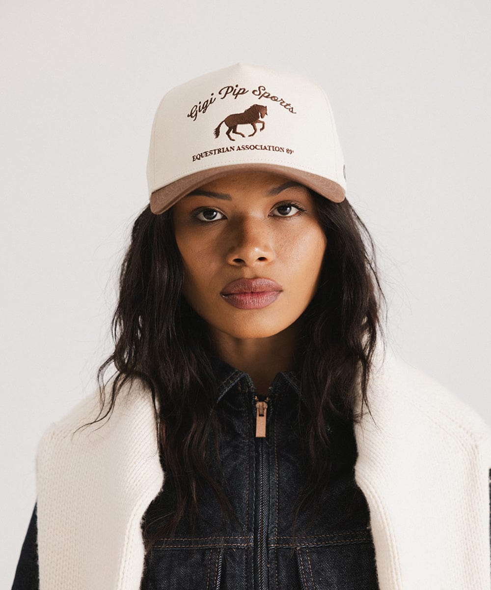Woman wearing a beige cap with text and a horse logo on a plain background