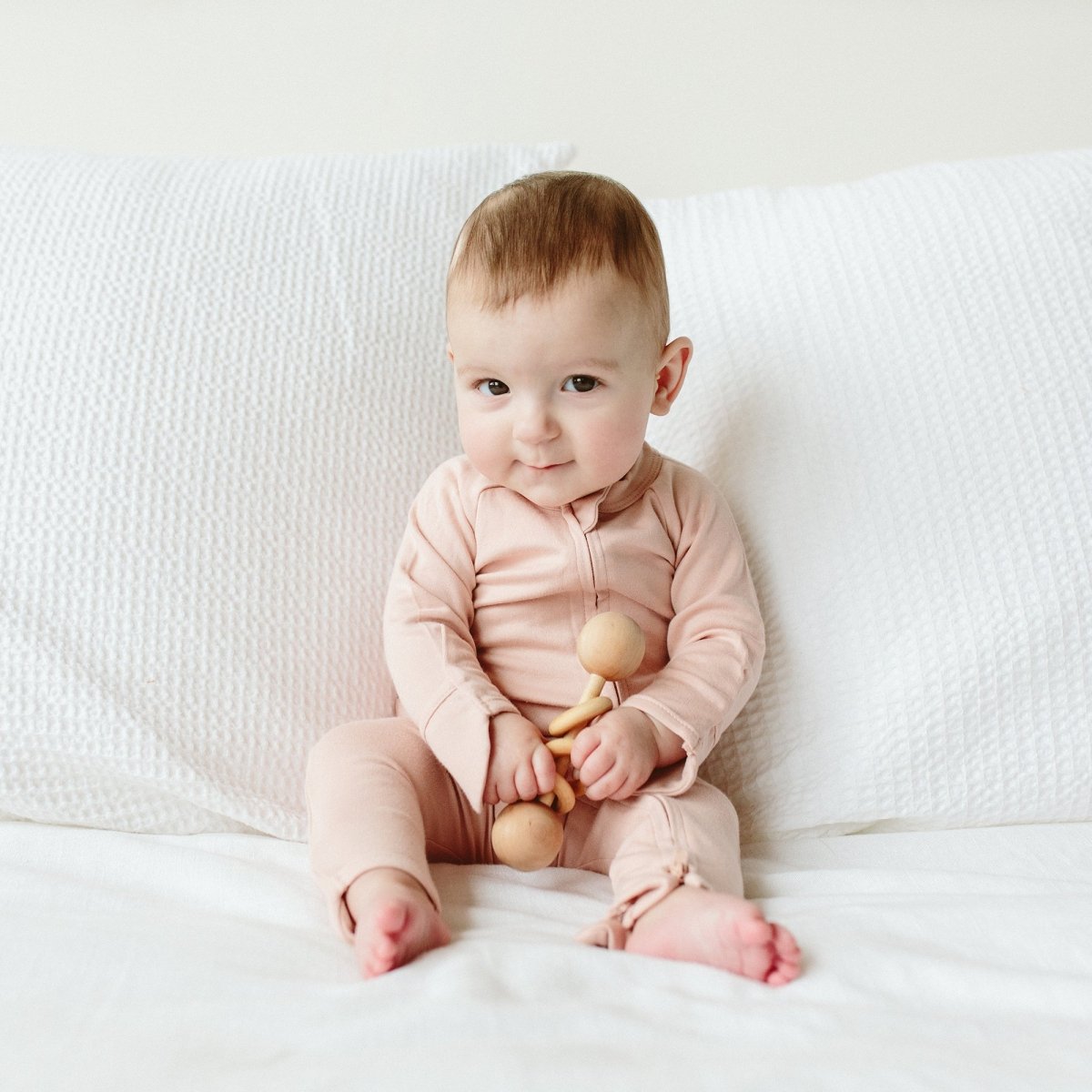 Baby in a pink outfit sitting on a white couch holding a wooden toy.