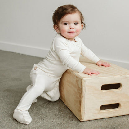 Baby in a white outfit playing with a wooden box on a carpeted floor.