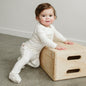 Baby in a white outfit playing with a wooden box on a carpeted floor.