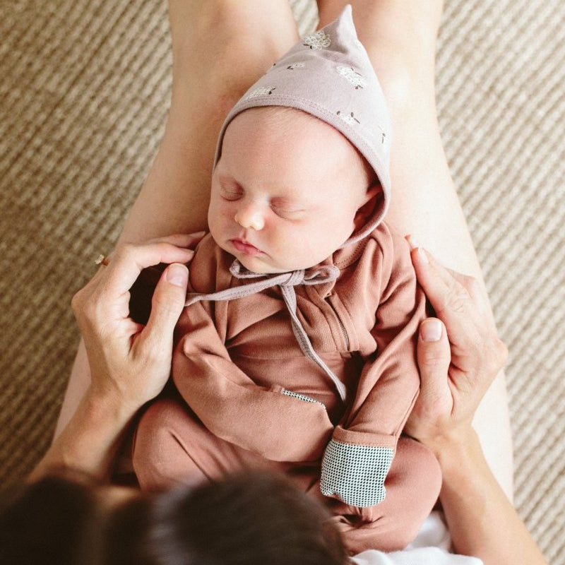 Newborn baby in pink outfit and hat being held by a person against a textured beige wall.