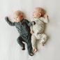 Two babies wearing gray and beige onesies lying on a white surface.