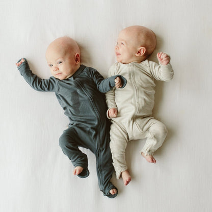 Two babies wearing gray and beige onesies lying on a white surface.