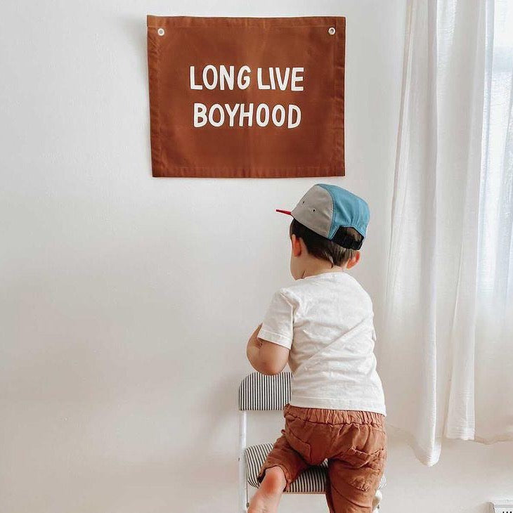 Child standing on a chair looking at a sign that says 'LONG LIVE BOYHOOD' on a white wall.