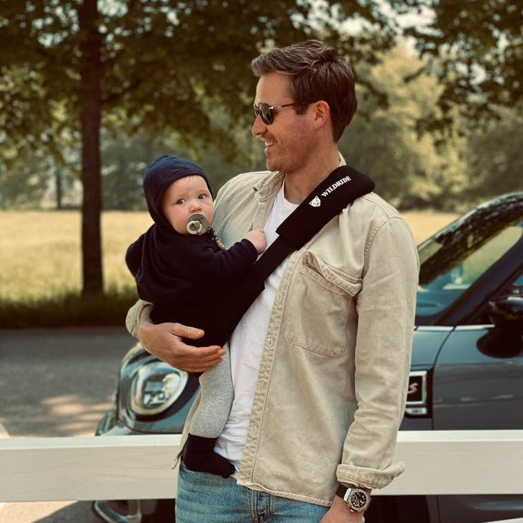 Man holding a baby in a sling outdoors with trees and a car in the background