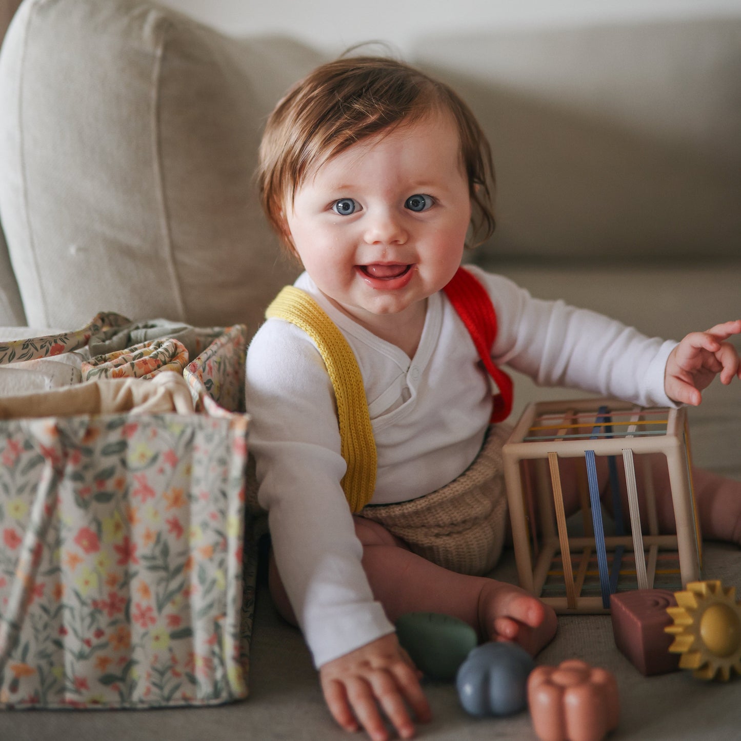 Baby sitting on a couch with toys around, wearing a white shirt and yellow suspenders.