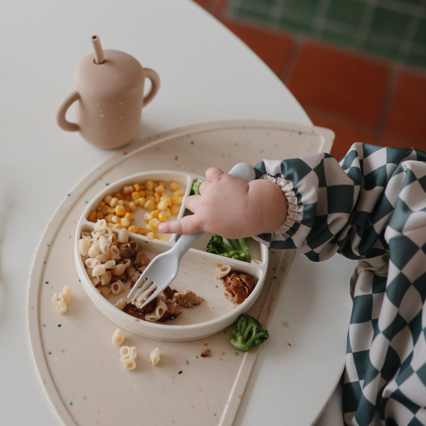 Child's hand reaching for food on a divided plate with a cup and tray in the background.