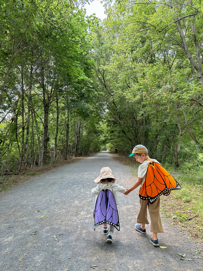 Two children in butterfly costumes walking on a path surrounded by trees