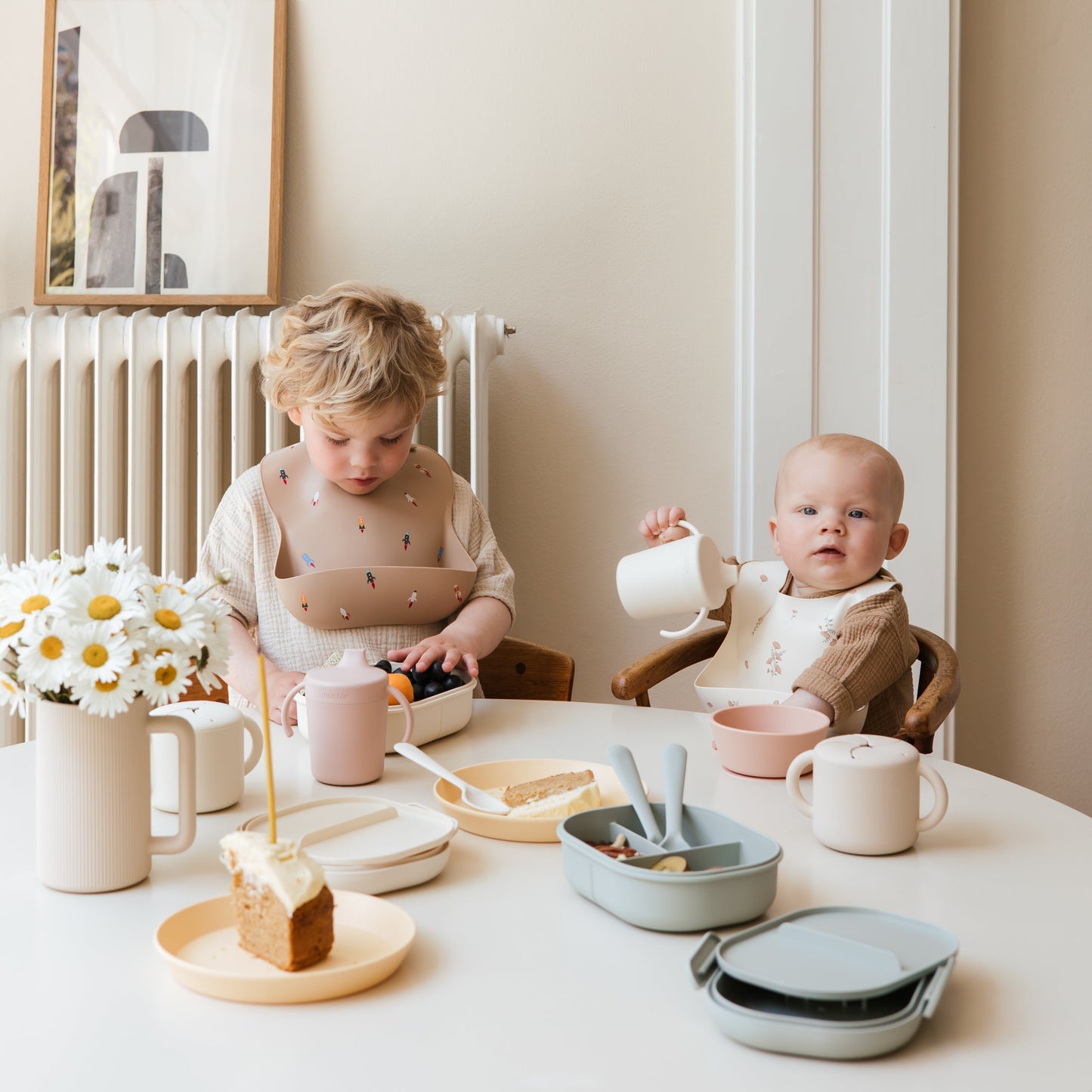 Two children sitting at a table with tableware and food, in a cozy indoor setting.