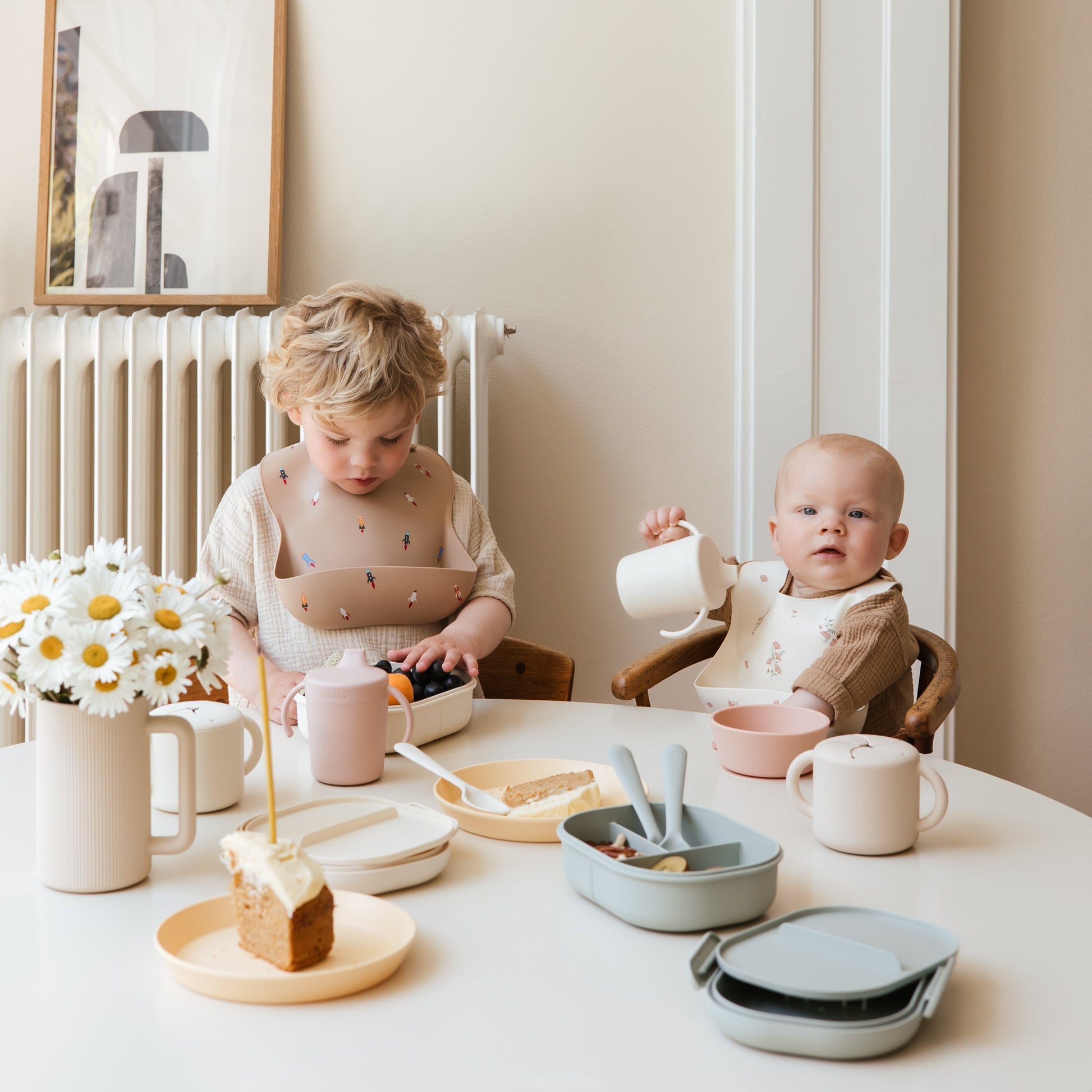 Two children sitting at a table with tableware and food, in a cozy indoor setting.