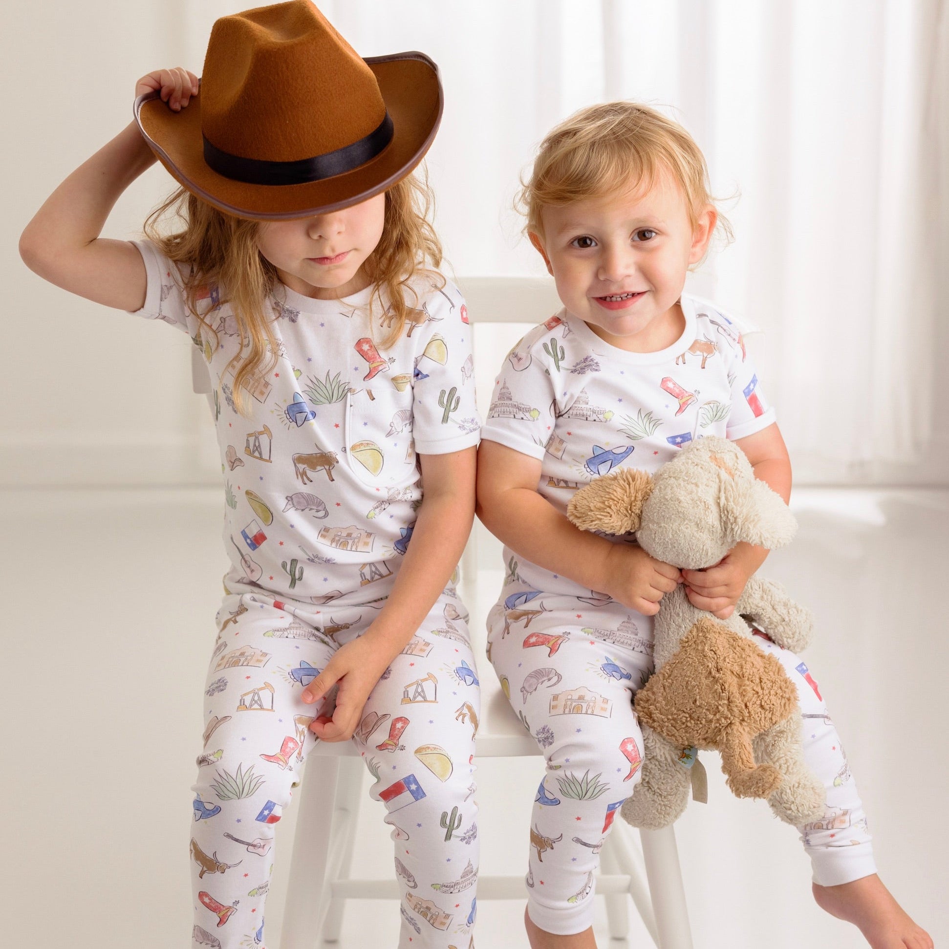 Two children in matching pajamas with animal prints, one wearing a hat and holding a teddy bear.