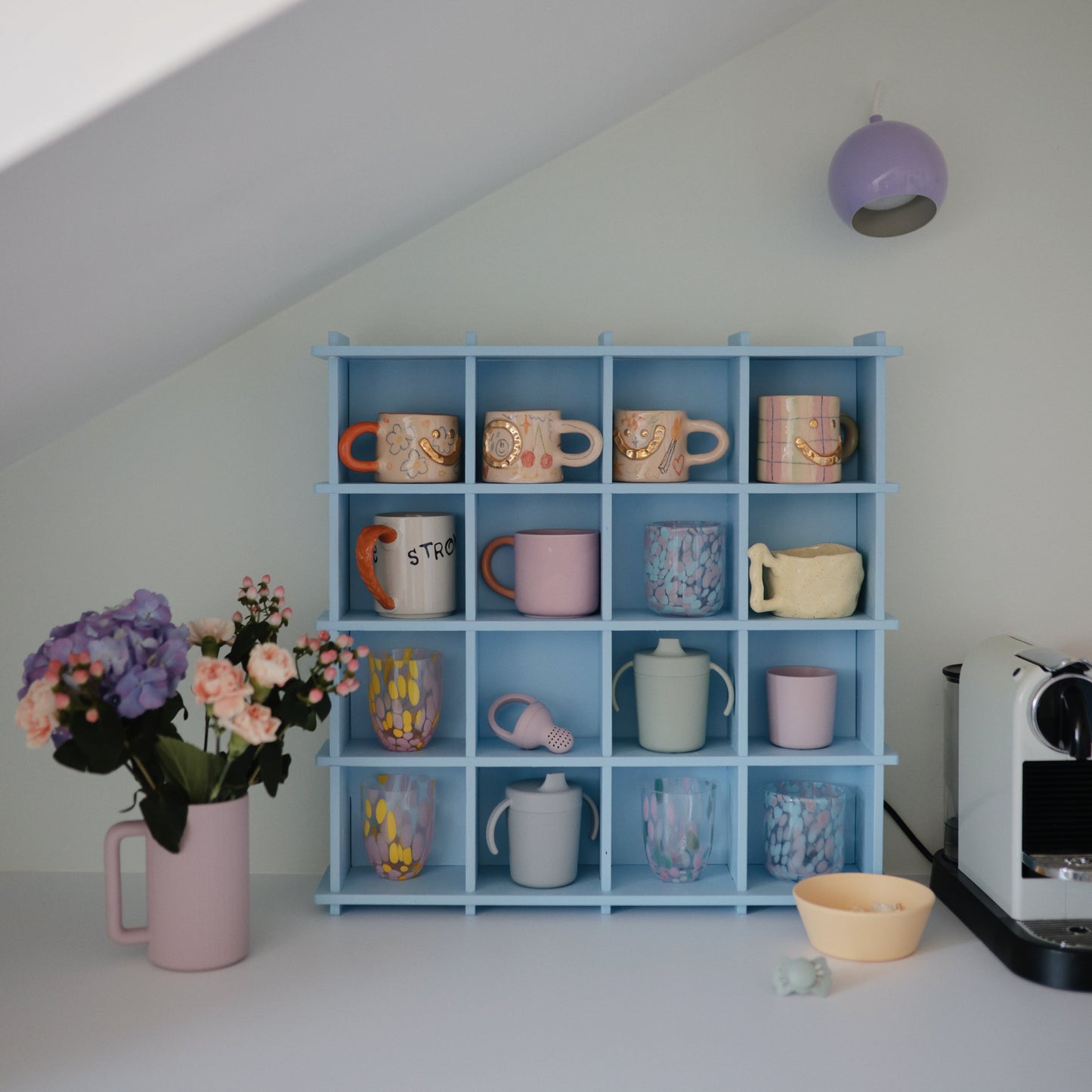 Collection of colorful mugs on a blue shelf with a pink vase and flowers in the foreground.