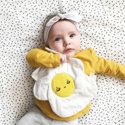 Baby wearing a yellow long-sleeve shirt and playing with a egg crinkle toy with a smiley face design on a patterned background.