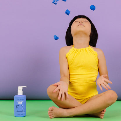 Child in yellow swimsuit sitting on a green floor with blue bottles and cubes against a purple background