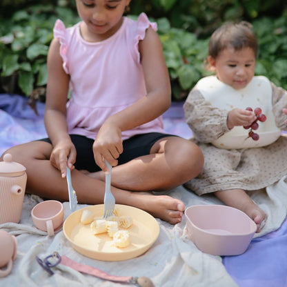 Two children sitting on a blanket outdoors with food and utensils.