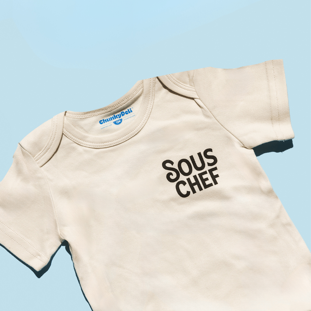 Beige baby onesie with 'Sous Chef' text on a light blue background