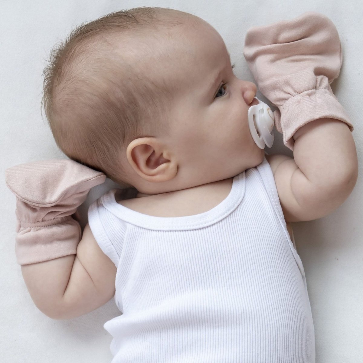 Baby lying on a white surface with pink gloves and a white pacifier.