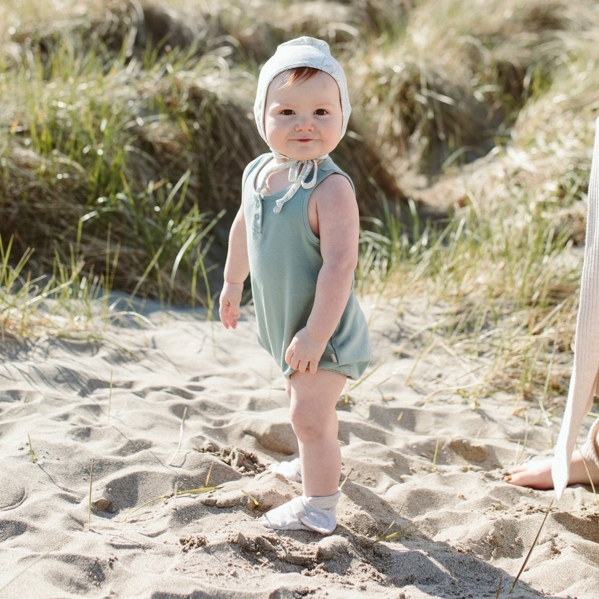 Baby in a light green outfit and white bonnet and light gray booties standing on sandy ground with grass in the background
