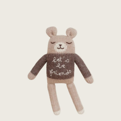 Knitted teddy bear wearing a sweater with 'let's be friends' text on a beige background