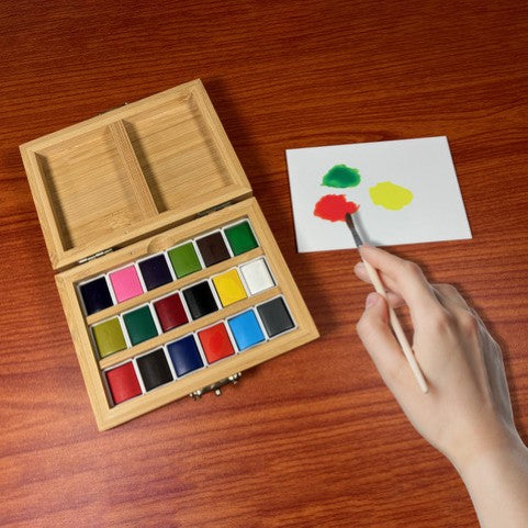 Wooden watercolor set with a hand holding a brush and a small painting on a wooden surface