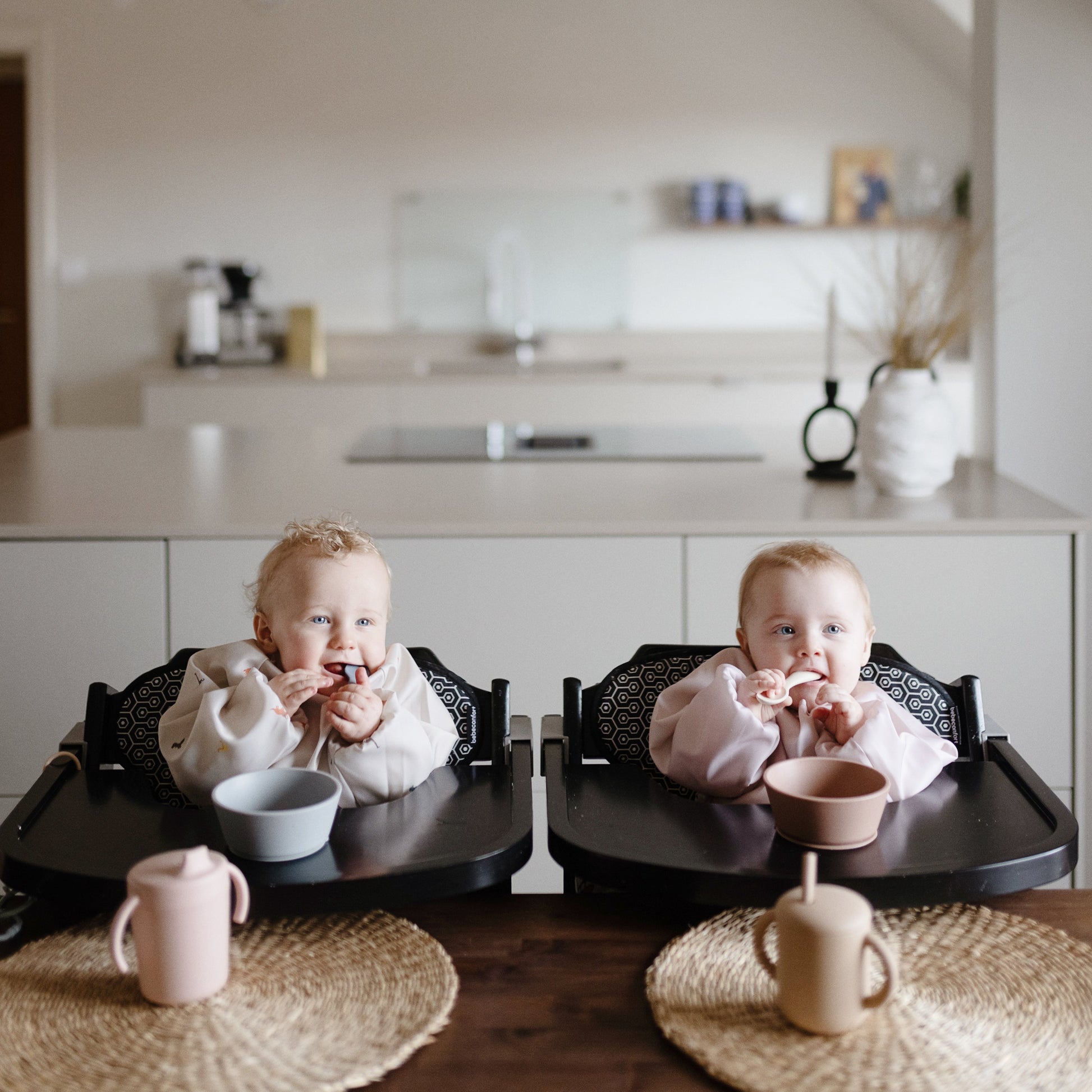 Two children sitting in high chairs in a kitchen eating.