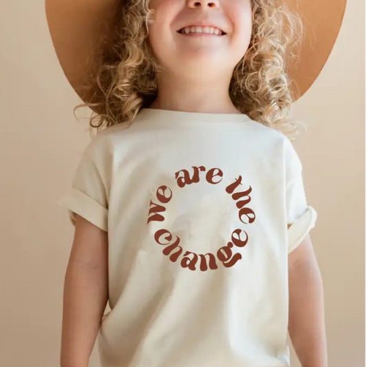 Child wearing a white t-shirt with 'we are the change' text, standing against a neutral background.