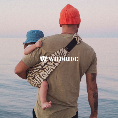 Man carrying a child on his back with a 'Wildride' bag, standing on a beach.