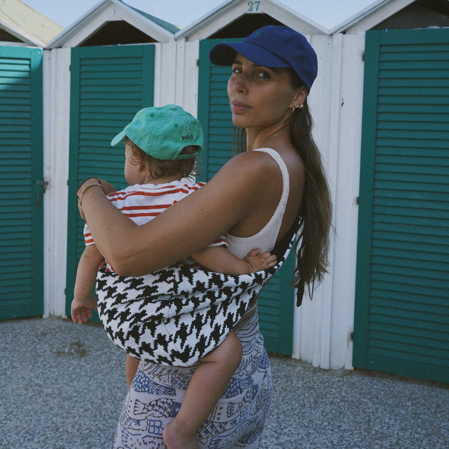 Woman holding a child in a houndstooth-patterned sling with beach huts in the background