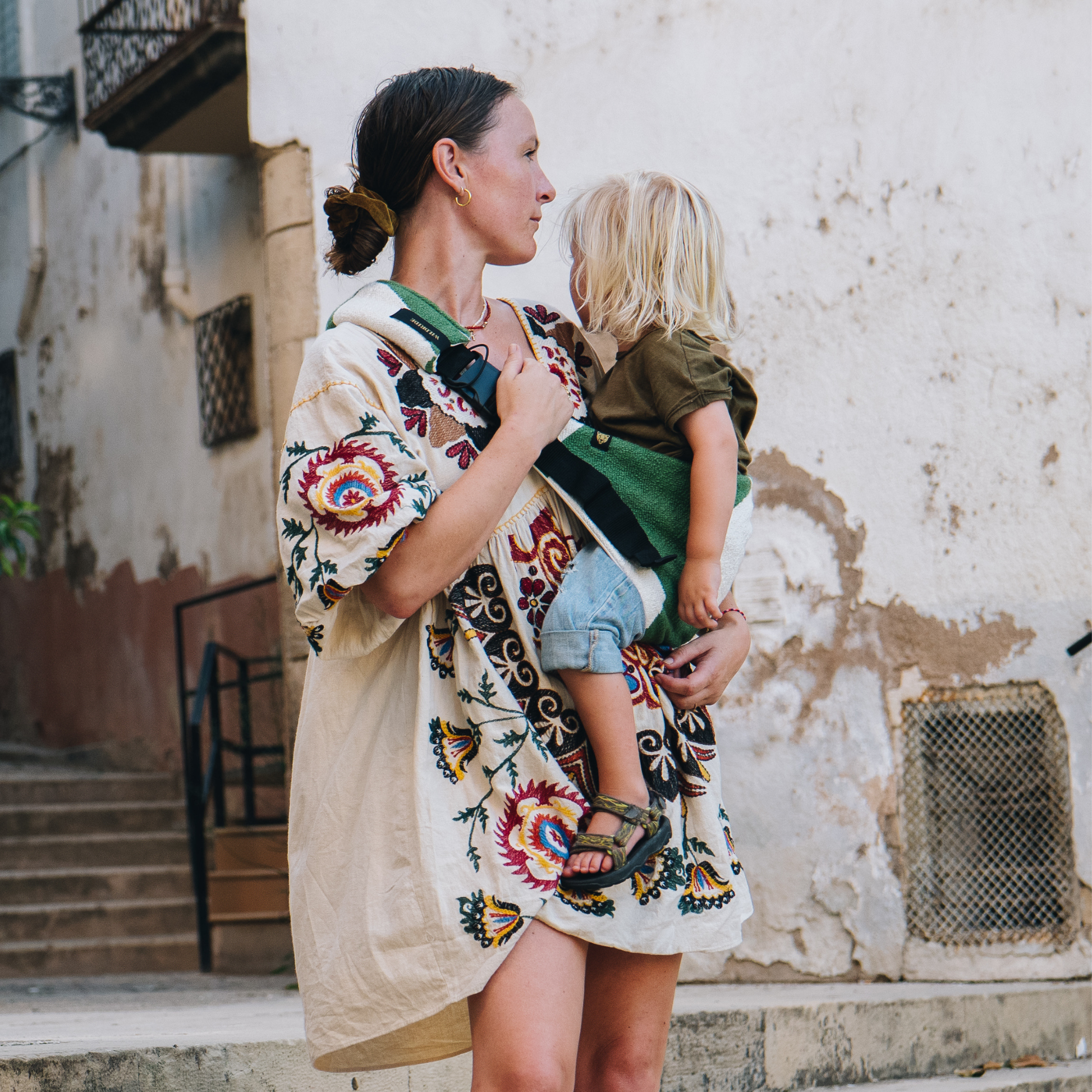 Woman carrying a child in a sling with a colorful floral pattern against a rustic wall.