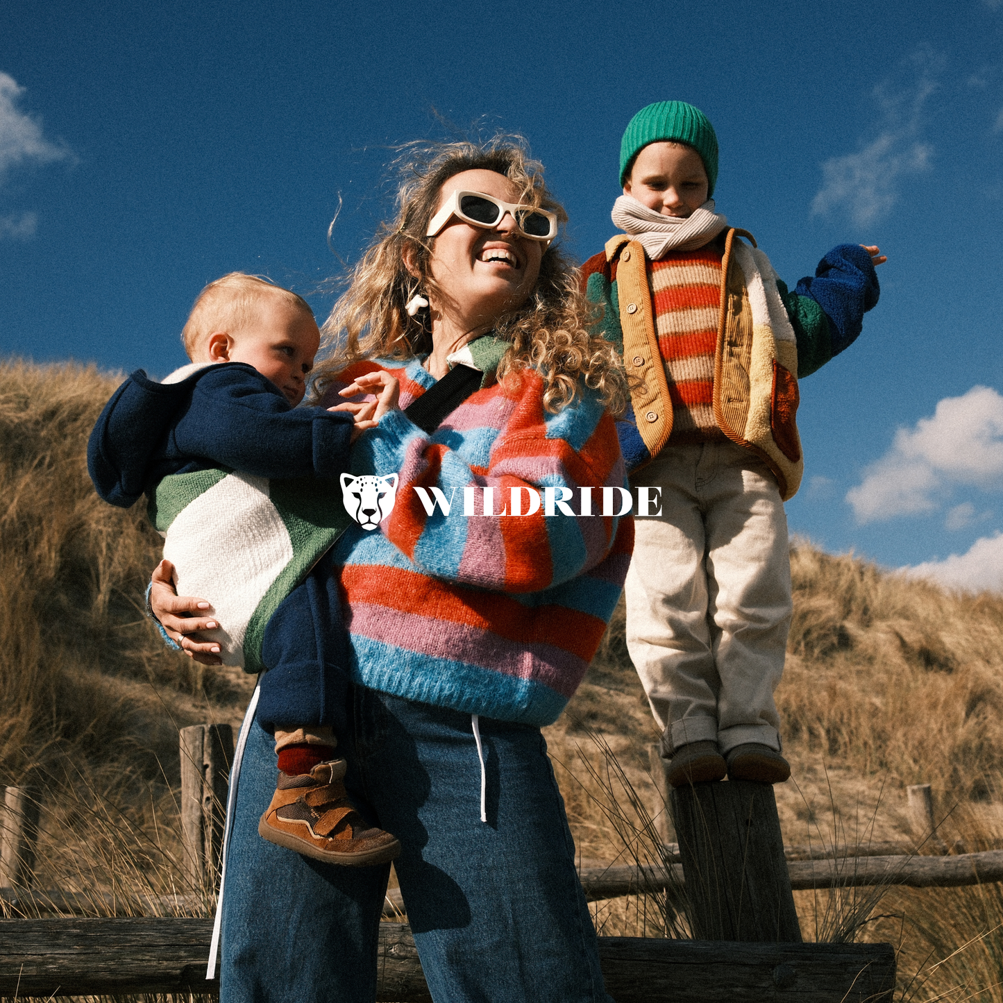 Woman holding a baby and standing next to a child on a wooden post with 'WILDRISE' branding.