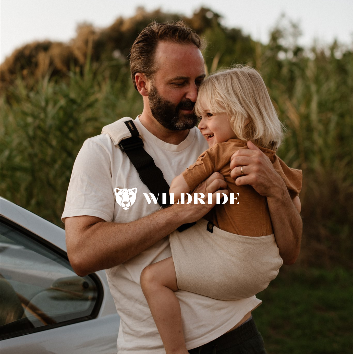 Man holding a child in a sling with 'Wildride' logo in a natural setting