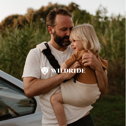Man holding a child in a sling with 'Wildride' logo in a natural setting