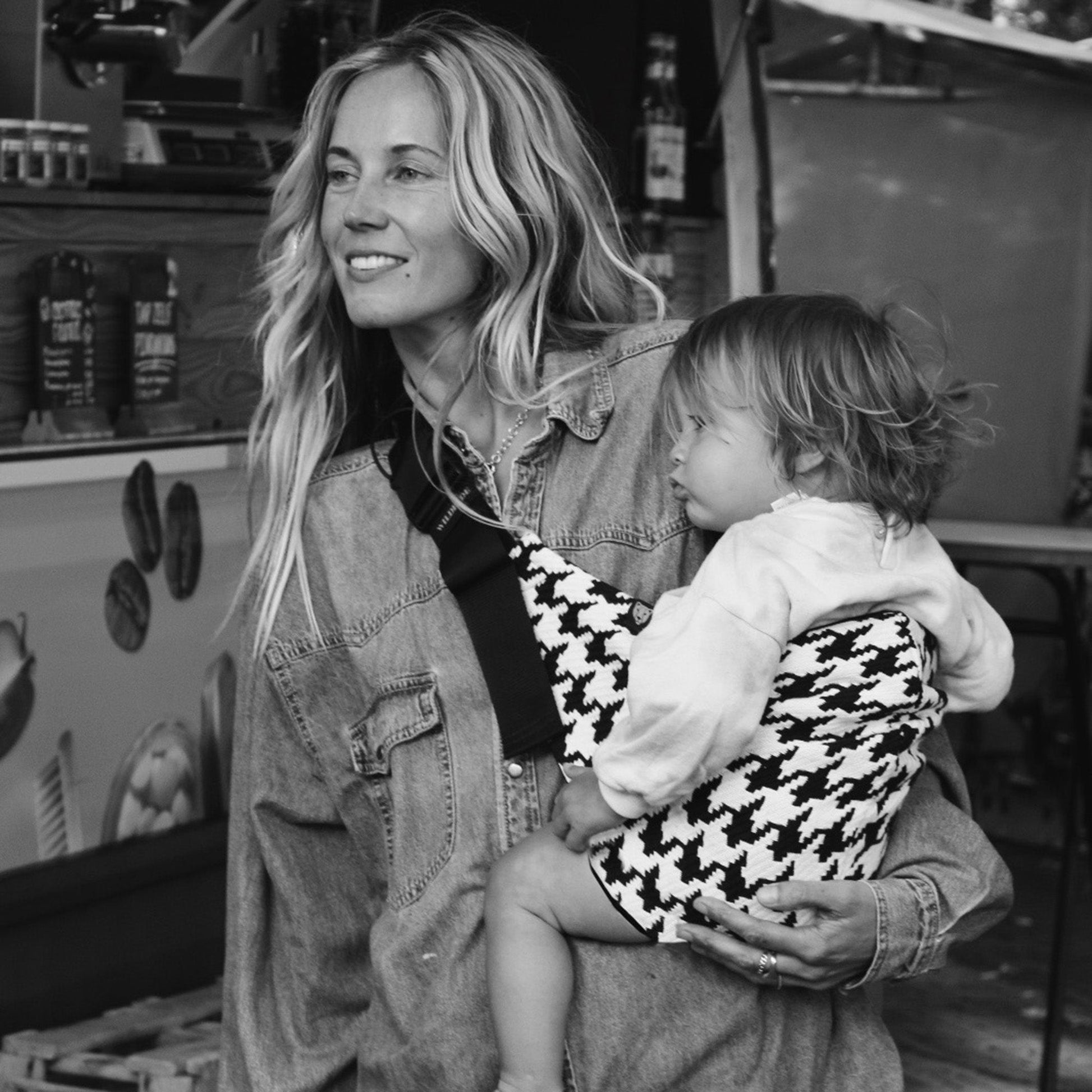 Woman holding a child in a houndstooth-patterned sling at an outdoor market.