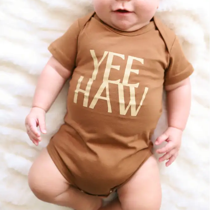 Baby wearing a brown onesie with 'YEE HAW' text on a white blanket